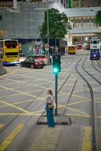 A woman in a blue dress stands at a green traffic light at a Hong Kong tram intersection, with yellow tram tracks, double-decker buses, cars, and a building with construction hoarding in the background