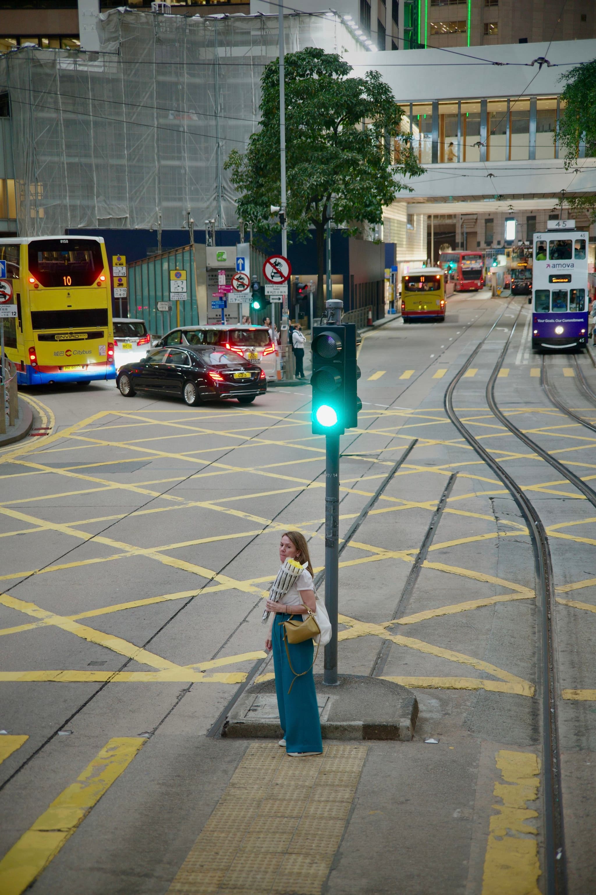 A woman in a blue dress stands at a green traffic light at a Hong Kong tram intersection, with yellow tram tracks, double-decker buses, cars, and a building with construction hoarding in the background