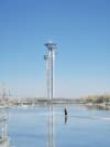 A solitary figure stands on a frozen expanse of ice, their reflection mirroring them on the smooth surface, set against the backdrop of the Beijing Olympic Tower piercing a clear blue sky