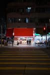 A night scene on a city street featuring a brightly lit shop with a vibrant red facade on the ground floor of a building In the foreground, a yellow pedestrian crosswalk spans the street, illuminated by streetlights. A red traffic light hangs above the intersection, while several people gather near the shop entrance and under a green awning next door